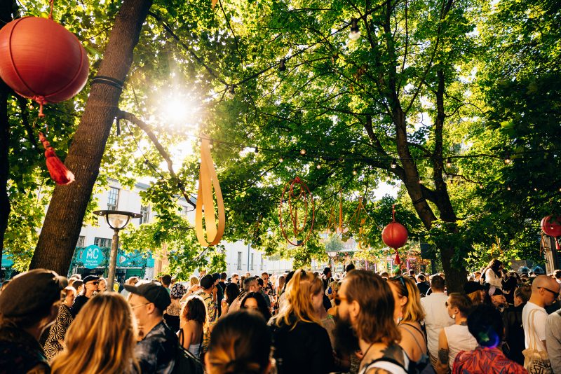 Ihmisiä puiden alla Flow Festivaaleilla kesällä 2023. Puista roikkuu erilaisia koristeita ja lamppuja.