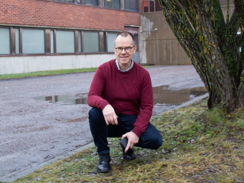 Mieshenkilö kyykistyneenä tiilisrakennuksen edessä poseeraa kameralle.