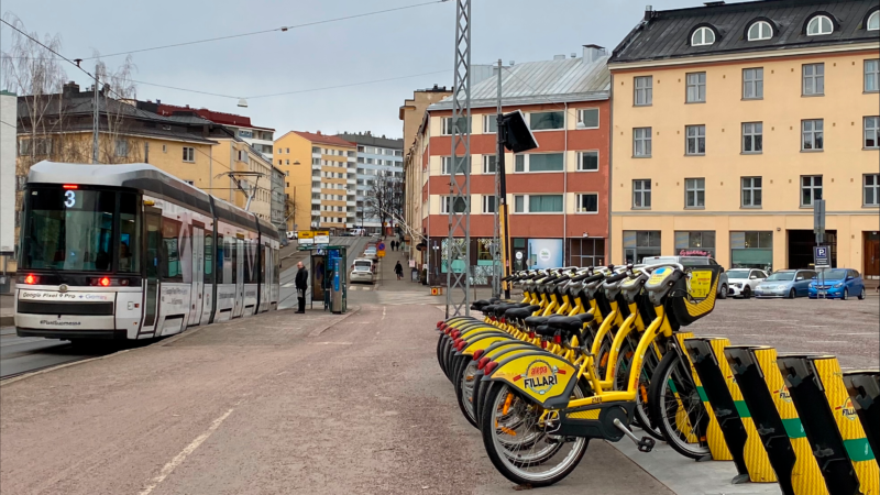 Kaupunkipyöriä Helsingin Alppilassa. Taustalla näkyy kerrostaloja. Pyörien vieressä on raitiovaunu.