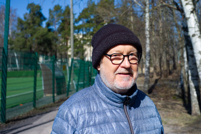 Vanhempi mieshenkilö sinisessä toppatakissa ja musta pipo päässä katsoo kameraan. Hänen taustalla siintää jalkapallokenttä.