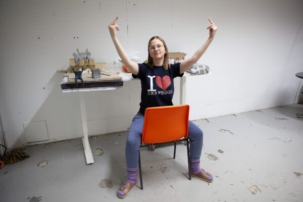 Laiho näyttää keskisormea molemmilla käsillään istuen tuolilla jalat leveillään. Hänellä on I LOVE DEAFHOOD -paita yllään. Laihon takana on pari keskisormea molemmilla käsillä näyttävää Kivikuuro- pienoisveistosta.