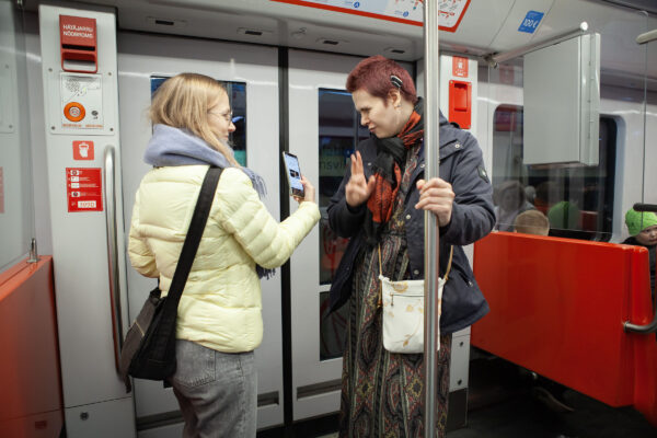 Metrossa Aino Laiho näyttää älypuhelintaan Lumo Kumpulaiselle. Lumolla on sisäkorvaistute. Aino ja Lumo seisovat metro-oven lähellä.