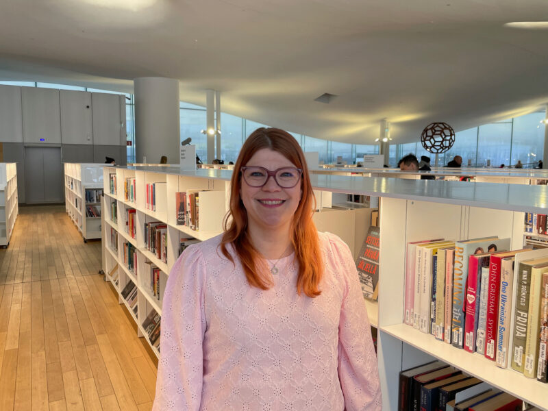 Nainen katsoo kohti kameraa. Taustalla kirjahyllyjä täynnä kirjoja.