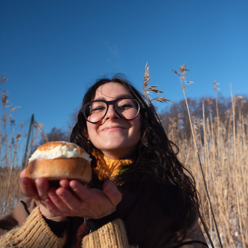 Jazzlaulaja pitelee kahdella kämmenellään laskiaispullaa leveä hymy huulillaan keskellä aurinkoista kaislikkoa.