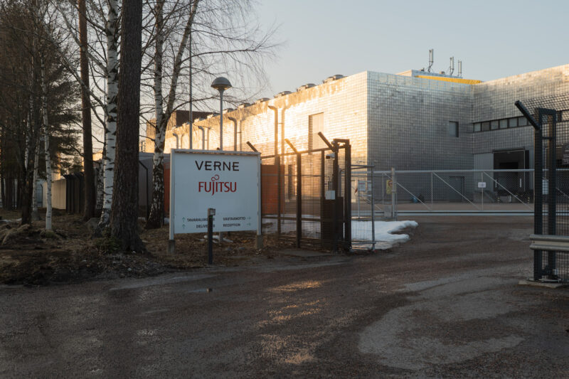 Datakeskustoimija Vernen datakeskus Vantaalla. Etualalla laitostoimijan kyltti ja avoin portti. Auringonlasku kimaltelee rakennuksen seinässä.