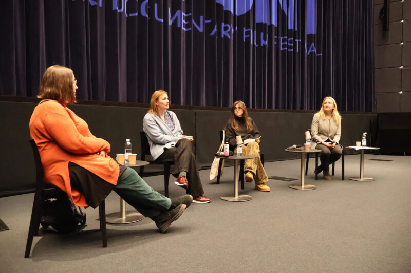 Panelistit istuvat yleisöä kohden väljästi rivissä, elokuvateatterin valkokankaan alla.