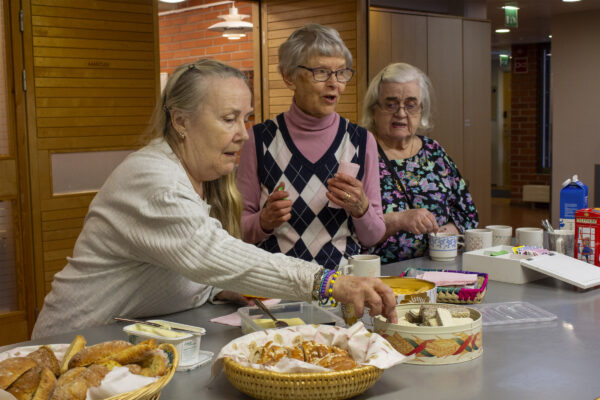 Kuvassa neulepiirin jäseniä ottamassa pöydästä kahvileipää. Pöydällä on tarjolla useita keksejä ja muuta syötävää.