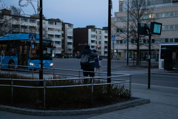 Bussikuski nousee seisomaan nojattuaan aitaan bussiasemalla. Vasemmalla sininen bussi.